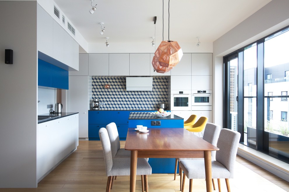 Moderne Wohnküche in L-Form mit flächenbündigen Schrankfronten, beigen Schränken, bunter Rückwand, Elektrogeräten mit Frontblende, hellem Holzboden und Kücheninsel in Sonstige