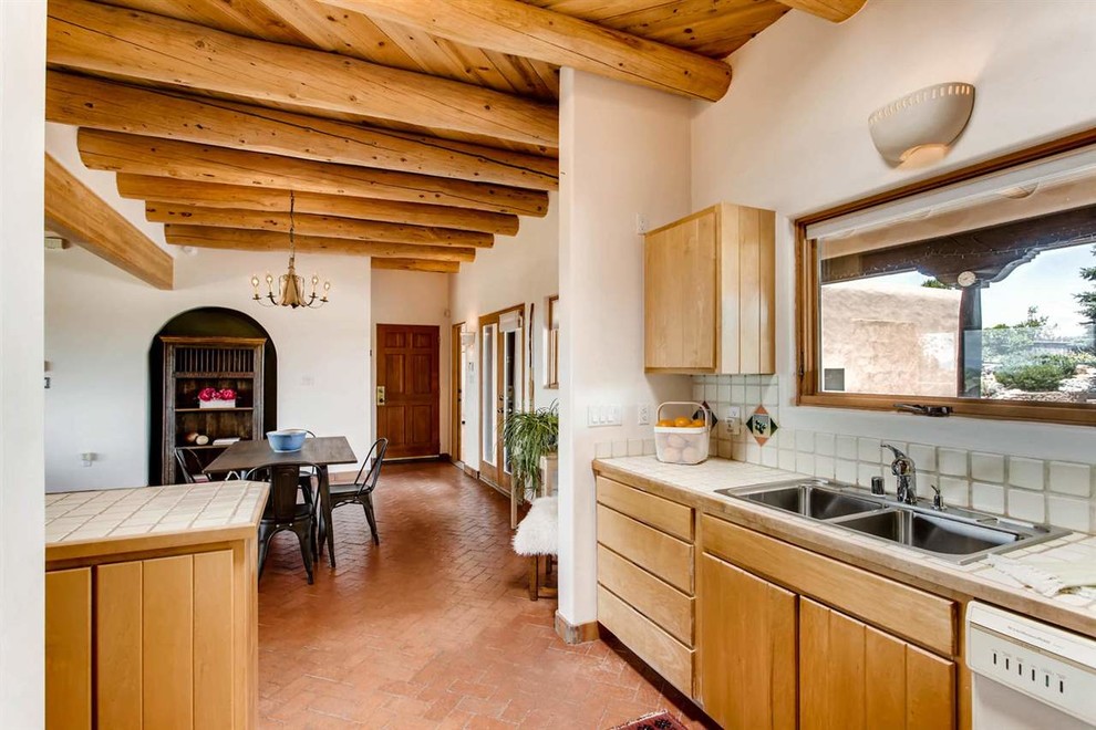 Foto på ett mellanstort amerikanskt kök, med en dubbel diskho, släta luckor, skåp i ljust trä, kaklad bänkskiva, vitt stänkskydd, stänkskydd i keramik, vita vitvaror, tegelgolv, en halv köksö och brunt golv