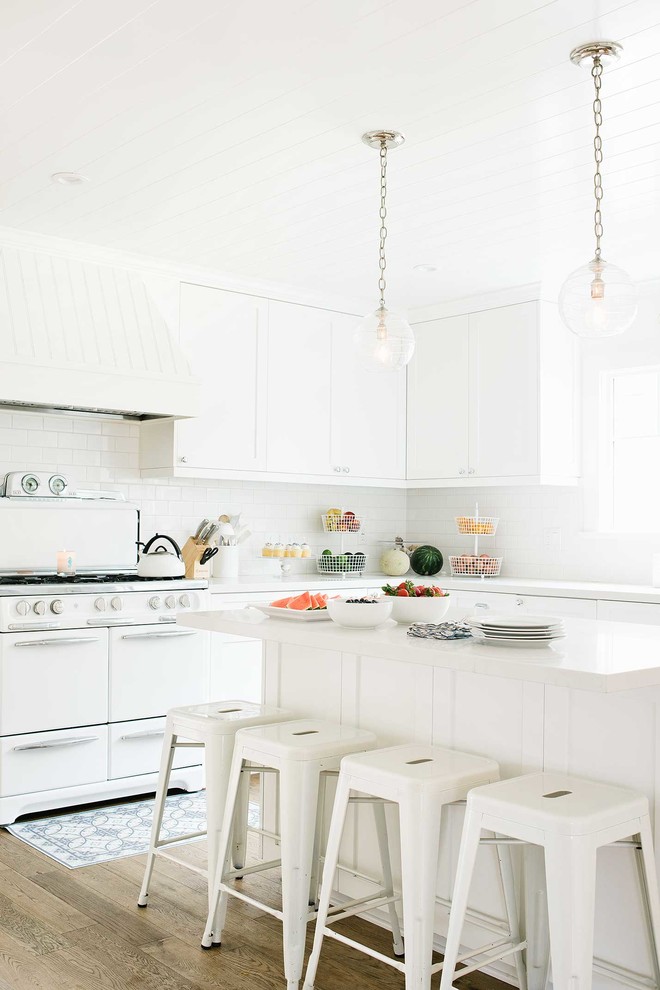 Photo of a coastal kitchen in Los Angeles with recessed-panel cabinets, white cabinets, white splashback, metro tiled splashback, white appliances, medium hardwood flooring and an island.