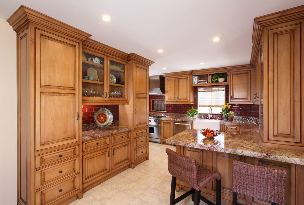 Foto på ett lantligt kök, med en rustik diskho, granitbänkskiva, rött stänkskydd, stänkskydd i glaskakel, rostfria vitvaror, luckor med upphöjd panel, skåp i mellenmörkt trä och klinkergolv i porslin