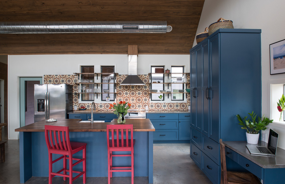 Lantlig inredning av ett kök, med en rustik diskho, skåp i shakerstil, blå skåp, träbänkskiva, flerfärgad stänkskydd, rostfria vitvaror, betonggolv och en köksö