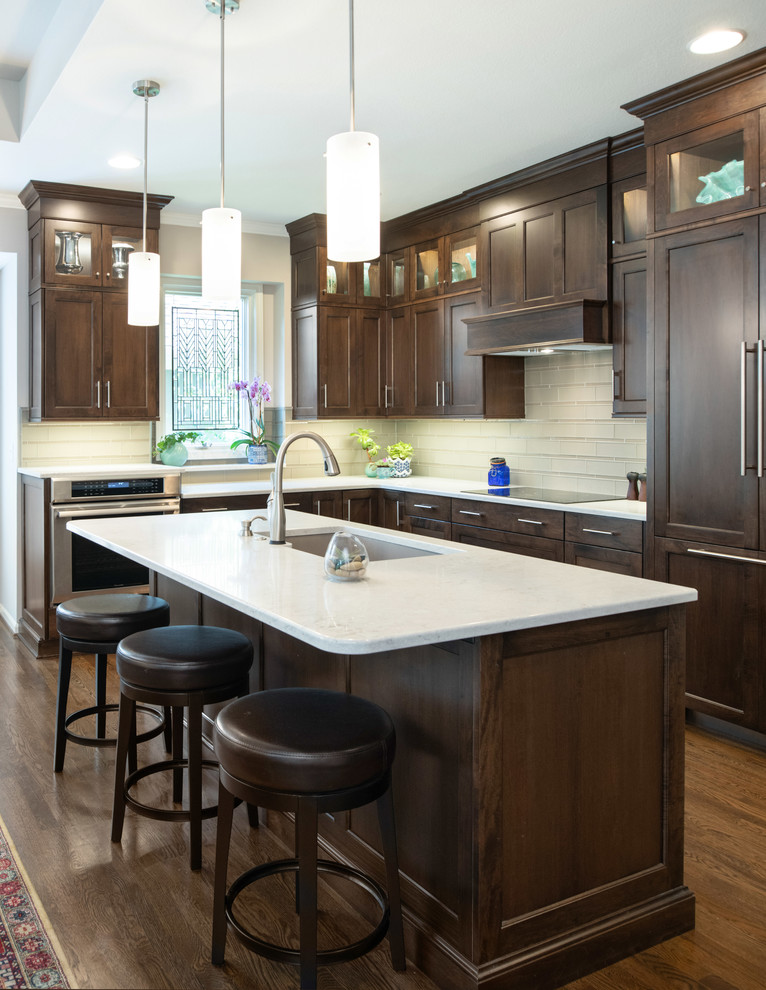 Medium sized traditional l-shaped open plan kitchen in Kansas City with shaker cabinets, dark wood cabinets, engineered stone countertops, glass tiled splashback, an island, a submerged sink, white splashback, stainless steel appliances, medium hardwood flooring, brown floors and white worktops.