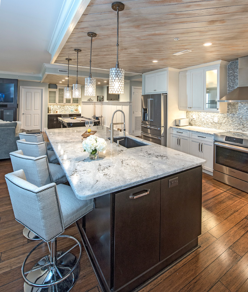 Inspiration for a mid-sized coastal single-wall medium tone wood floor open concept kitchen remodel in Other with a drop-in sink, flat-panel cabinets, white cabinets, marble countertops, gray backsplash, glass tile backsplash, stainless steel appliances and an island