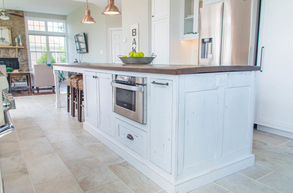 This is an example of a medium sized farmhouse galley kitchen/diner in DC Metro with a belfast sink, flat-panel cabinets, white cabinets, wood worktops, white splashback, stainless steel appliances, porcelain flooring, an island, beige floors and brown worktops.