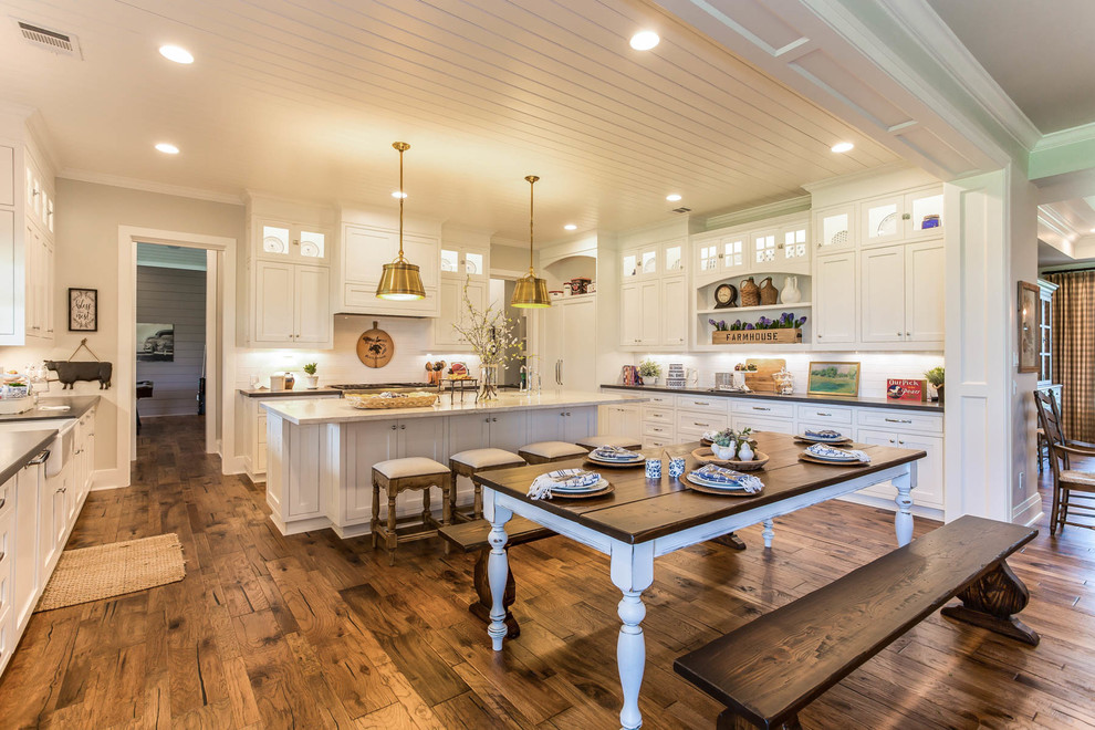 Cette image montre une cuisine américaine rustique avec un évier de ferme, des portes de placard blanches, une crédence blanche, un électroménager blanc, un sol en bois brun, îlot et un sol marron.
