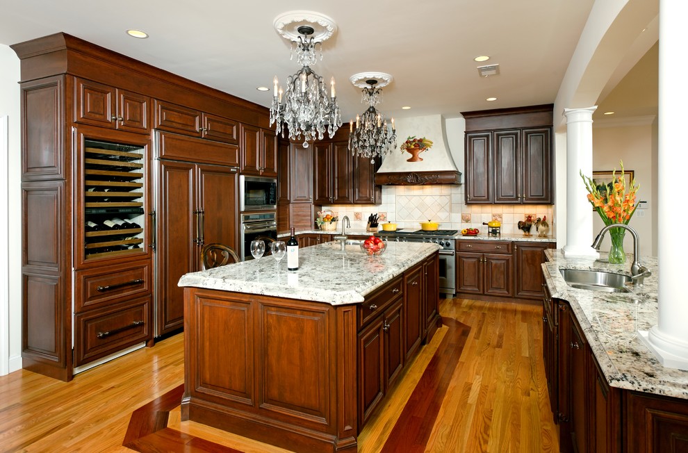 Example of a classic u-shaped light wood floor eat-in kitchen design in DC Metro with an undermount sink, raised-panel cabinets, dark wood cabinets, granite countertops, white backsplash, stone tile backsplash, stainless steel appliances and two islands