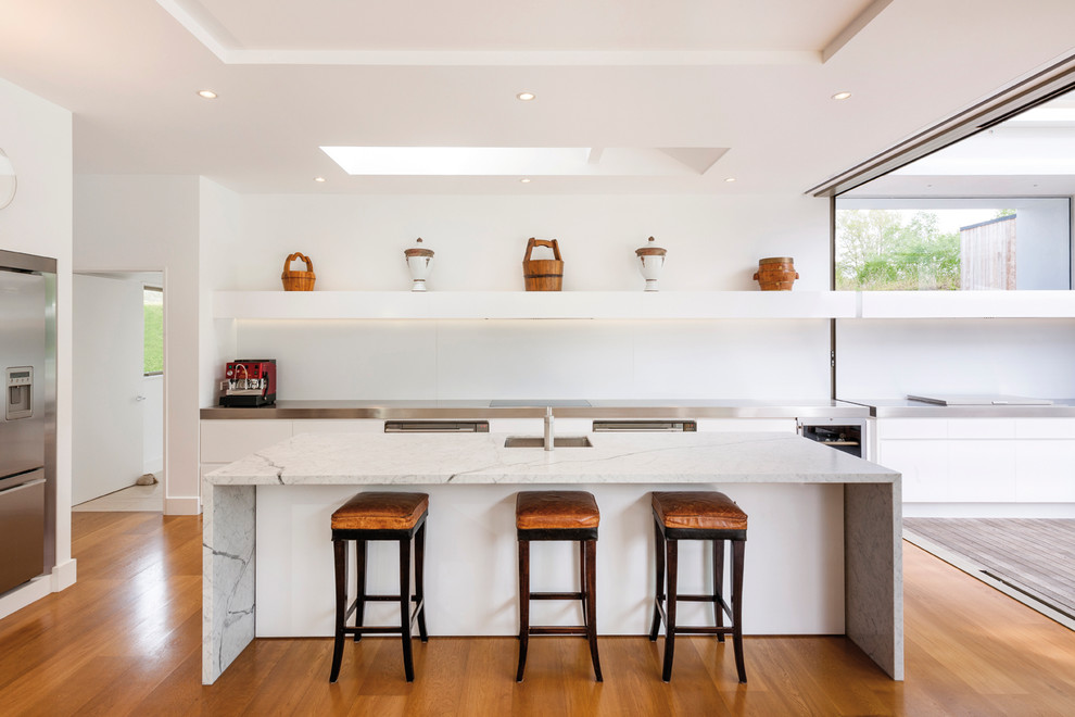 Moderne Küche in L-Form mit flächenbündigen Schrankfronten, weißen Schränken, Küchenrückwand in Weiß, Küchengeräten aus Edelstahl, braunem Holzboden und Kücheninsel in Sonstige
