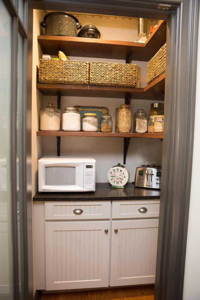 This is an example of a small victorian galley kitchen pantry in Chicago with a belfast sink, recessed-panel cabinets, distressed cabinets, granite worktops, beige splashback, ceramic splashback, black appliances, light hardwood flooring, no island, brown floors and black worktops.