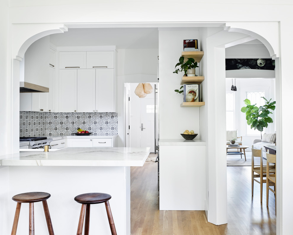 Photo of a medium sized scandinavian u-shaped enclosed kitchen in San Francisco with shaker cabinets, white cabinets, marble worktops, multi-coloured splashback, cement tile splashback, stainless steel appliances, light hardwood flooring, a breakfast bar, brown floors and white worktops.