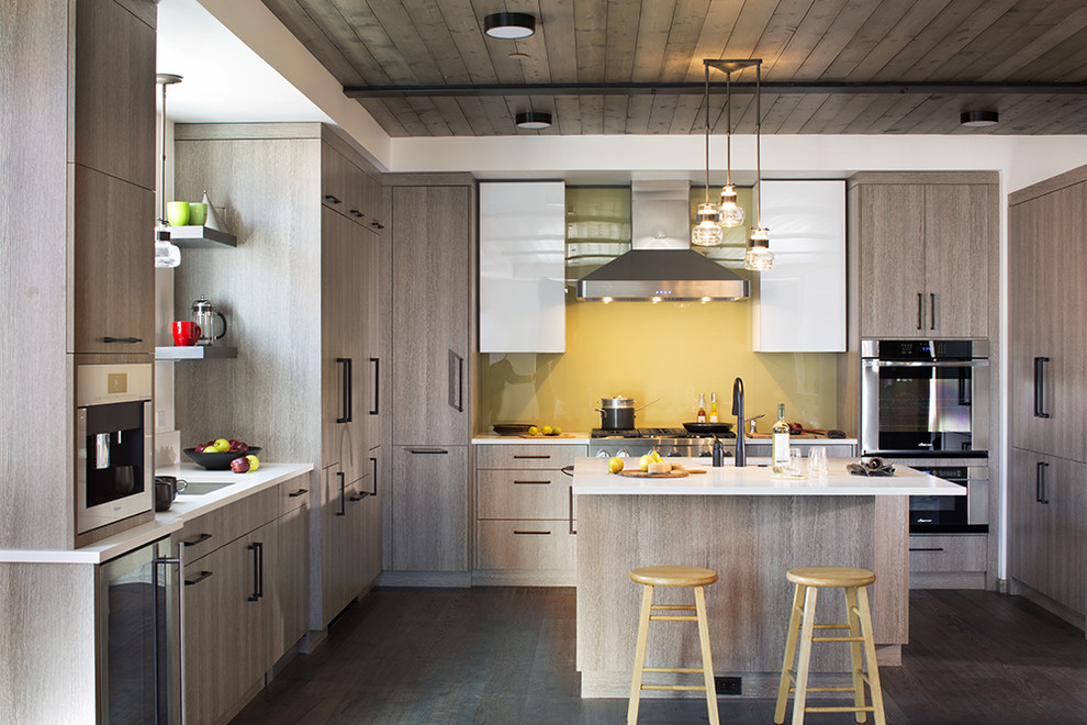 Bild på ett funkis vit vitt kök, med en undermonterad diskho, släta luckor, skåp i mellenmörkt trä, gult stänkskydd, glaspanel som stänkskydd, rostfria vitvaror, mörkt trägolv, en köksö och brunt golv