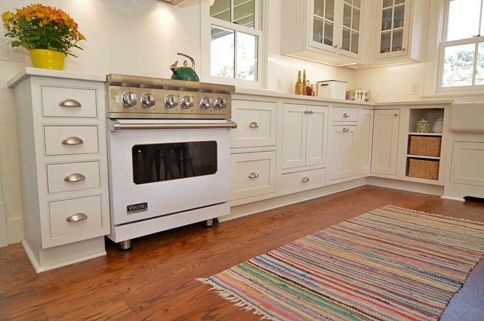 Photo of a small country u-shaped kitchen/diner in Other with a belfast sink, white appliances, beaded cabinets, white cabinets, engineered stone countertops, white splashback, medium hardwood flooring and no island.