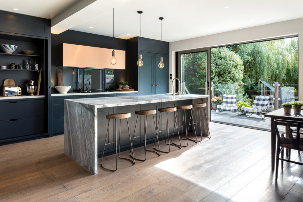 Design ideas for a medium sized contemporary galley kitchen/diner in London with a built-in sink, flat-panel cabinets, blue cabinets, marble worktops, blue splashback, glass sheet splashback, black appliances, medium hardwood flooring, an island, brown floors and grey worktops.