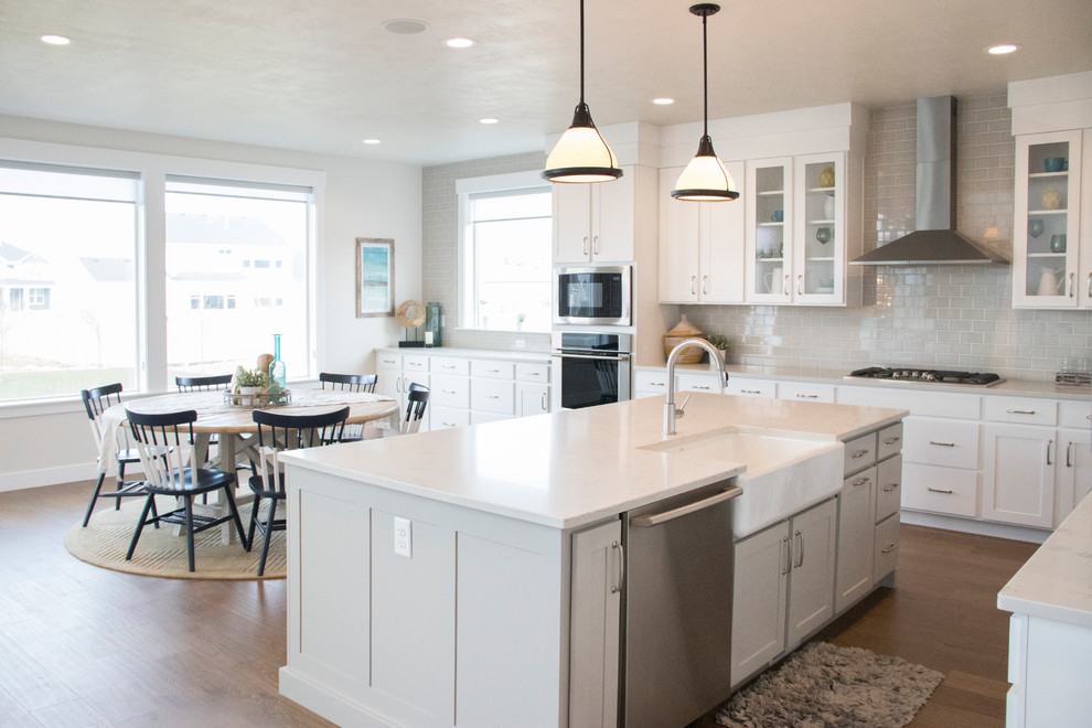This is an example of a large traditional l-shaped kitchen/diner in Salt Lake City with a belfast sink, shaker cabinets, white cabinets, engineered stone countertops, grey splashback, ceramic splashback, stainless steel appliances, medium hardwood flooring, an island and brown floors.