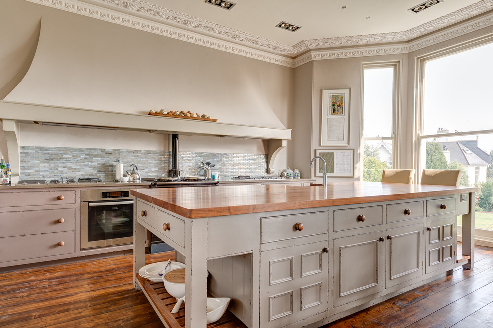 Design ideas for a large victorian kitchen/diner in Devon with a double-bowl sink, recessed-panel cabinets, distressed cabinets, wood worktops, grey splashback, ceramic splashback, stainless steel appliances, medium hardwood flooring and an island.