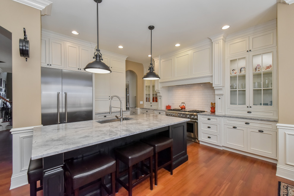 This is an example of a medium sized traditional l-shaped kitchen in Chicago with recessed-panel cabinets, white cabinets, engineered stone countertops, white splashback, ceramic splashback, stainless steel appliances, medium hardwood flooring, an island, brown floors and grey worktops.