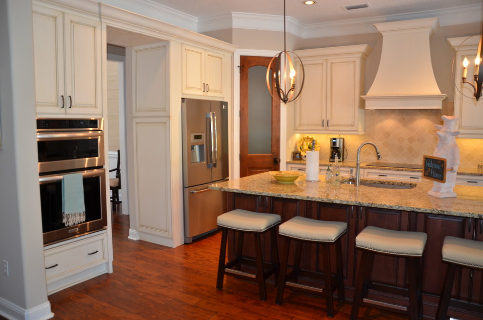 Example of a classic l-shaped medium tone wood floor kitchen design in Jacksonville with an undermount sink, raised-panel cabinets, white cabinets, granite countertops, beige backsplash, stone tile backsplash, stainless steel appliances and an island