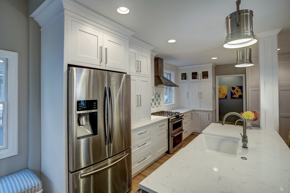 Design ideas for a medium sized classic galley kitchen/diner in Grand Rapids with a belfast sink, shaker cabinets, white cabinets, engineered stone countertops, white splashback, ceramic splashback, stainless steel appliances, light hardwood flooring and an island.