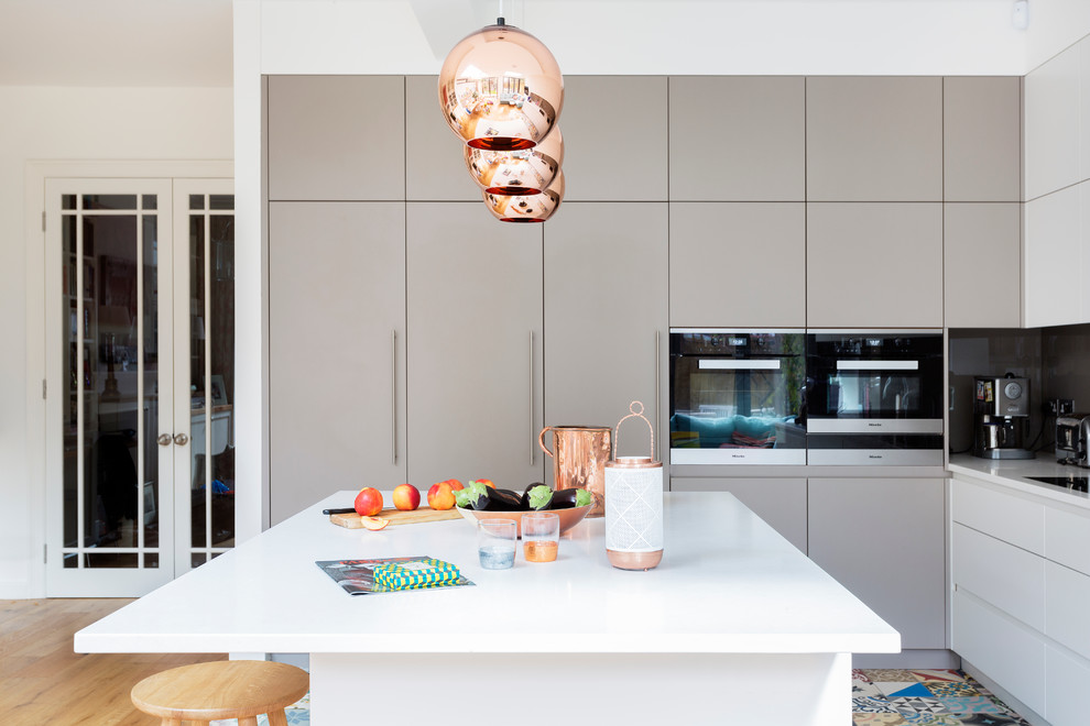 Photo of a contemporary l-shaped kitchen in London with flat-panel cabinets, white cabinets, grey splashback, glass sheet splashback, light hardwood flooring and an island.