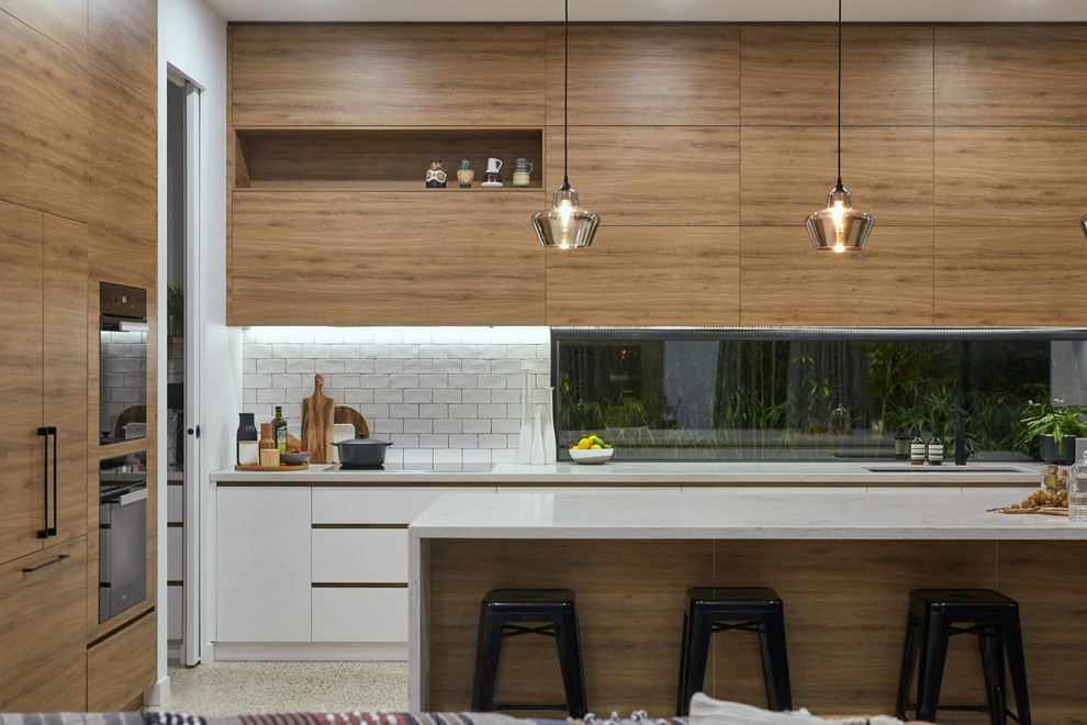 Moderne Küche in L-Form mit Unterbauwaschbecken, flächenbündigen Schrankfronten, hellbraunen Holzschränken, Küchenrückwand in Weiß, Rückwand-Fenster, Elektrogeräten mit Frontblende, Betonboden, Kücheninsel, grauem Boden und grauer Arbeitsplatte in Melbourne