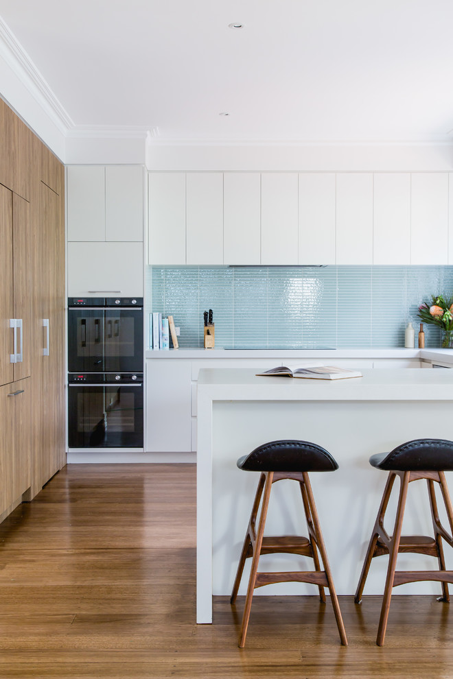Foto de cocina minimalista de tamaño medio con despensa, puertas de armario de madera clara, salpicadero azul, salpicadero de azulejos de vidrio, electrodomésticos negros y suelo marrón