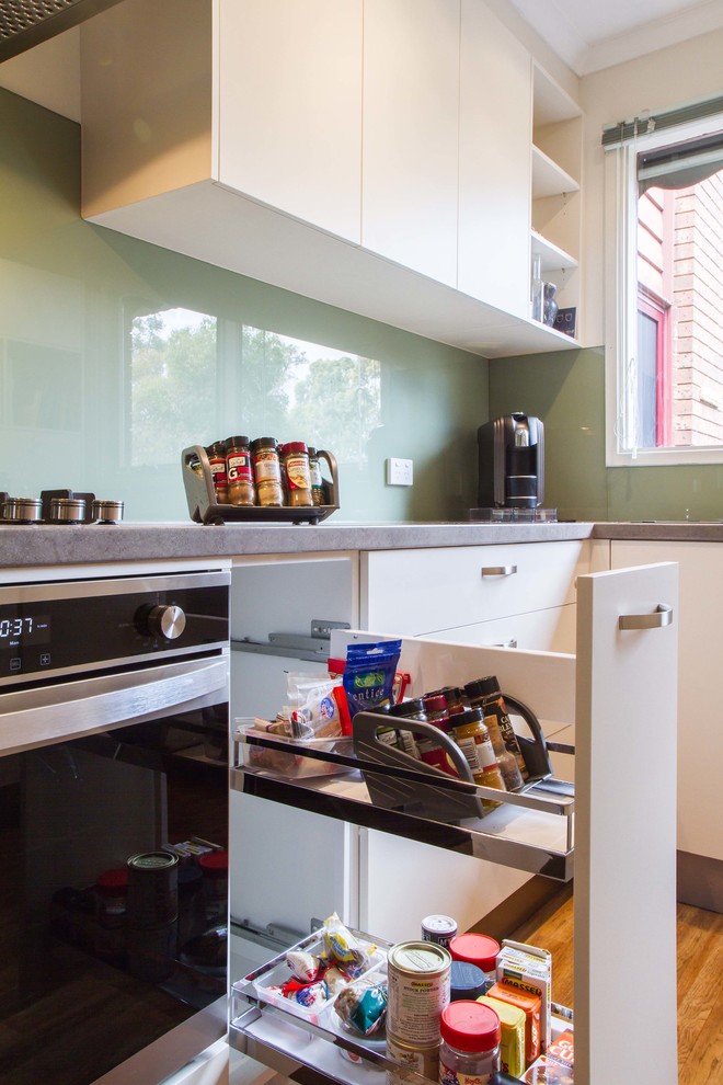 Geschlossene, Kleine Moderne Küche ohne Insel in U-Form mit Einbauwaschbecken, flächenbündigen Schrankfronten, weißen Schränken, Laminat-Arbeitsplatte, Küchenrückwand in Grün, Glasrückwand und braunem Holzboden in Melbourne