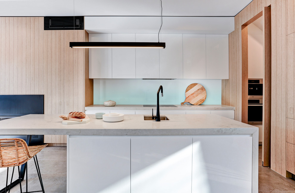 Photo of a large contemporary galley open plan kitchen in Melbourne with a submerged sink, white cabinets, engineered stone countertops, white splashback, glass sheet splashback, black appliances, porcelain flooring, flat-panel cabinets, an island, brown floors and grey worktops.