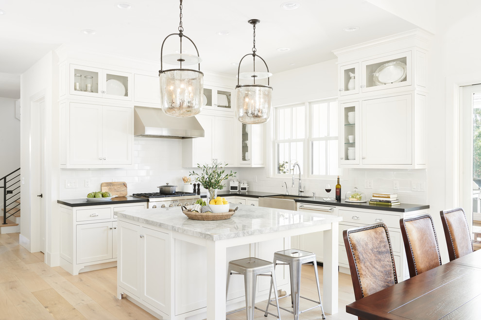 Inspiration for a country l-shaped open plan kitchen in San Francisco with a belfast sink, shaker cabinets, white cabinets, white splashback, metro tiled splashback, stainless steel appliances, light hardwood flooring, an island, black worktops and beige floors.