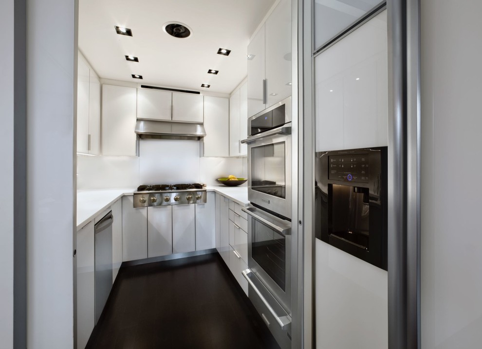 Photo of a small modern u-shaped kitchen/diner in New York with a submerged sink, flat-panel cabinets, white cabinets, engineered stone countertops, white splashback, stone slab splashback, stainless steel appliances, dark hardwood flooring, a breakfast bar, brown floors and white worktops.