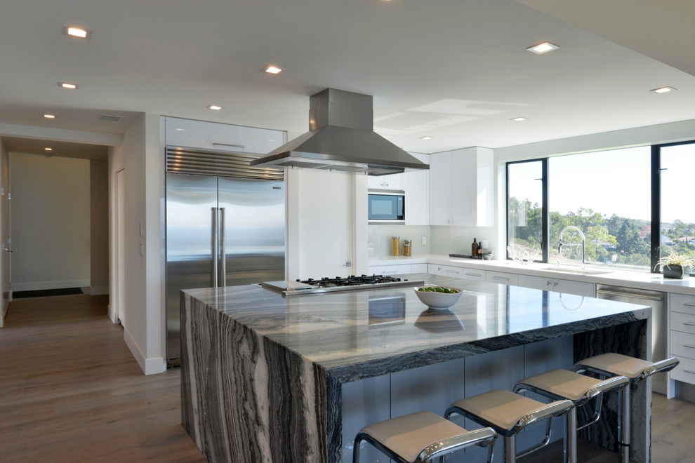 Example of a large minimalist galley light wood floor eat-in kitchen design in San Diego with an undermount sink, flat-panel cabinets, white cabinets, marble countertops, white backsplash, glass sheet backsplash, stainless steel appliances and an island