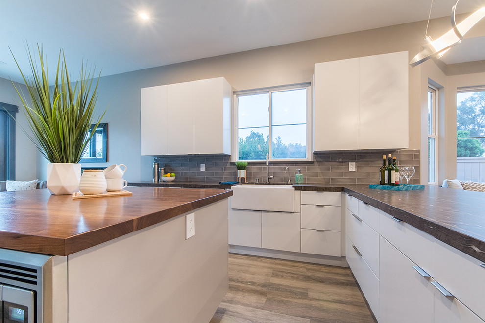 Photo of a medium sized contemporary galley open plan kitchen in San Diego with a belfast sink, flat-panel cabinets, white cabinets, engineered stone countertops, grey splashback, metro tiled splashback, stainless steel appliances, laminate floors, an island, brown floors and grey worktops.