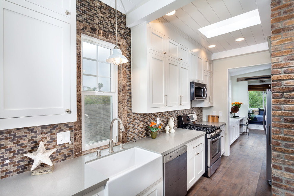 Cette image montre une cuisine américaine parallèle design avec un évier de ferme, un placard à porte shaker, des portes de placard blanches, une crédence marron et un électroménager en acier inoxydable.