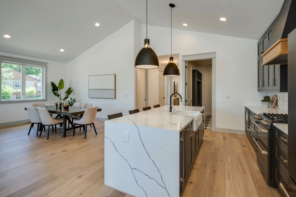 Example of a mid-sized transitional galley light wood floor and vaulted ceiling eat-in kitchen design in Denver with a farmhouse sink, shaker cabinets, gray cabinets, quartz countertops, white backsplash, ceramic backsplash, black appliances, an island and white countertops