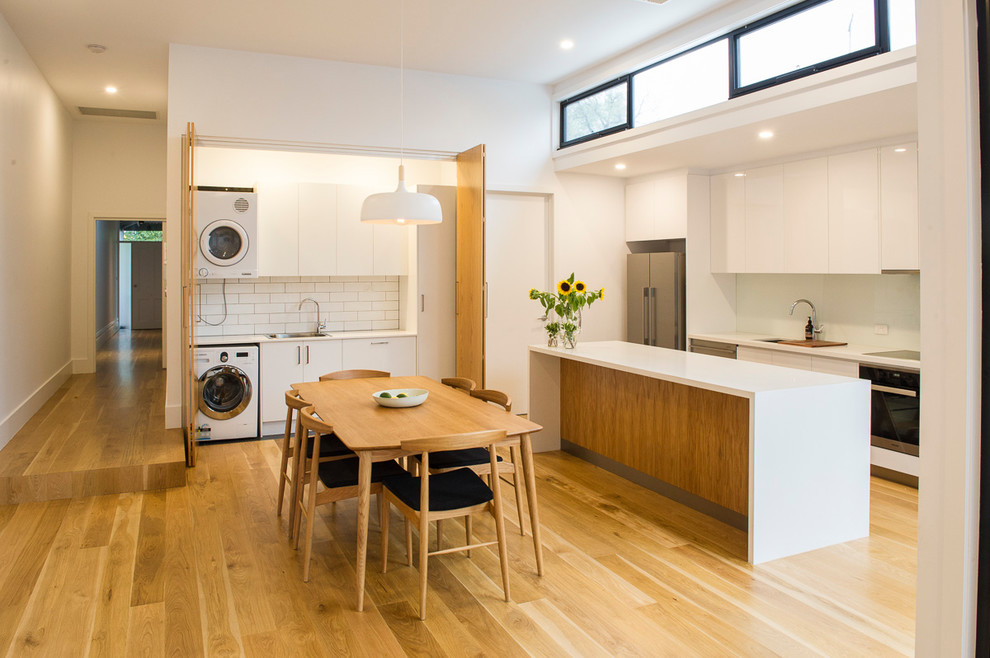 Modern inredning av ett kök, med släta luckor, vita skåp, vitt stänkskydd, stänkskydd i tunnelbanekakel, rostfria vitvaror, mellanmörkt trägolv, en köksö och brunt golv
