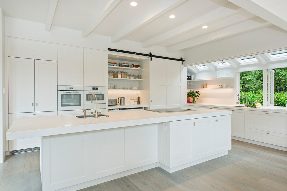 Inspiration for a large timeless l-shaped light wood floor and beige floor open concept kitchen remodel in Auckland with an undermount sink, shaker cabinets, white cabinets, quartz countertops, white backsplash, white appliances and an island
