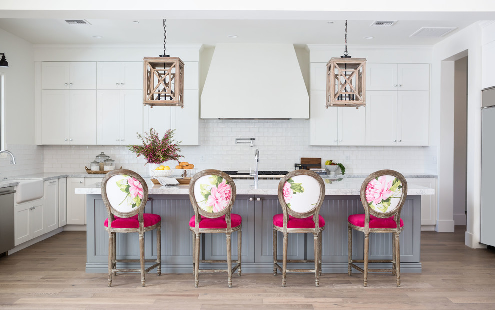 Photo of a farmhouse grey and pink l-shaped kitchen in Phoenix with a belfast sink, shaker cabinets, white cabinets, white splashback, metro tiled splashback, stainless steel appliances, light hardwood flooring, an island, beige floors and grey worktops.