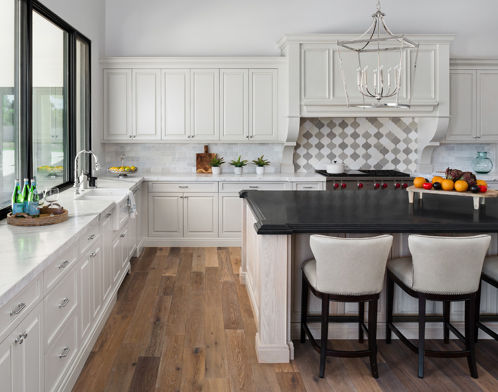 Inspiration for a transitional l-shaped medium tone wood floor and brown floor kitchen remodel in Phoenix with a farmhouse sink, raised-panel cabinets, white cabinets, multicolored backsplash, stainless steel appliances, an island and white countertops