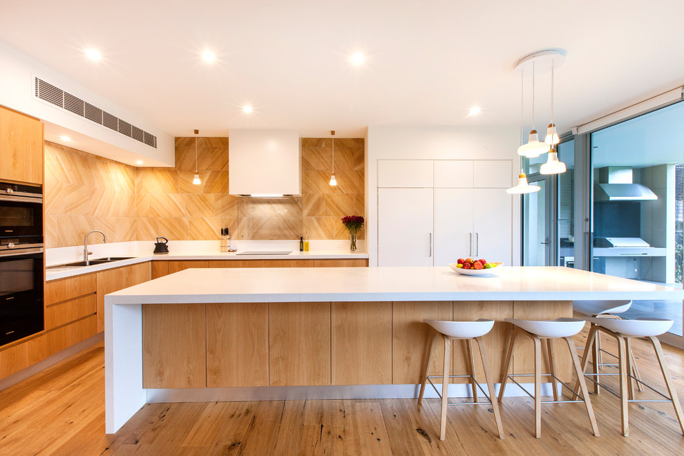Large trendy l-shaped medium tone wood floor open concept kitchen photo in Adelaide with light wood cabinets, solid surface countertops, an island, a double-bowl sink, flat-panel cabinets, brown backsplash and black appliances