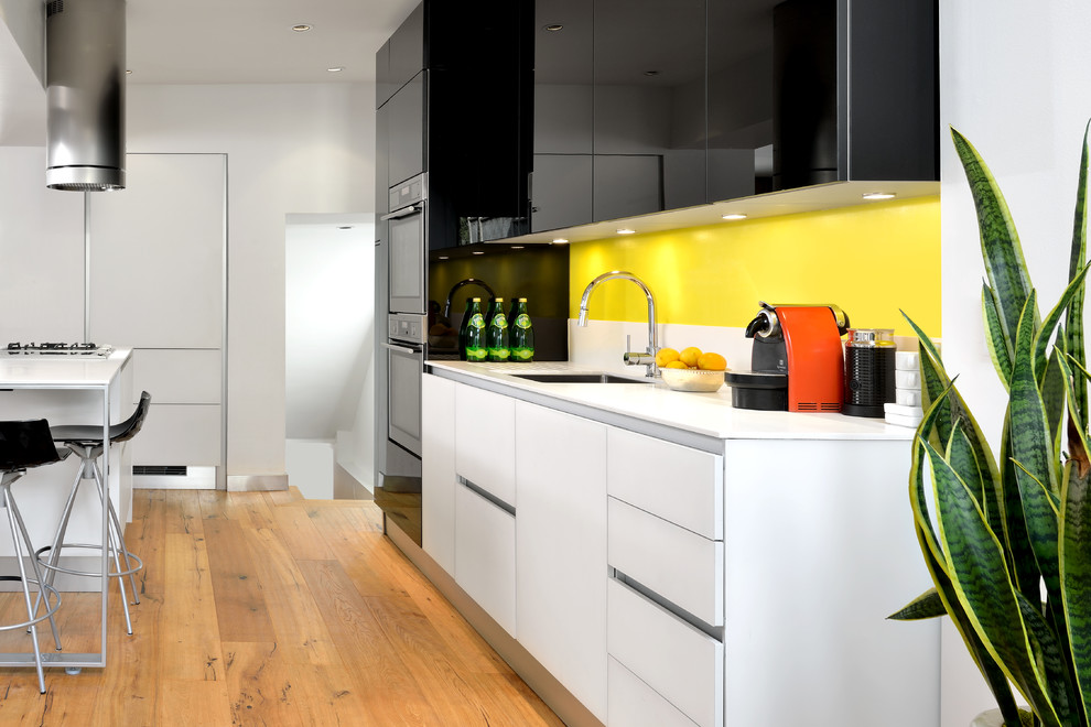 На фото: параллельная кухня-гостиная в стиле модернизм с врезной мойкой, плоскими фасадами, черными фасадами, столешницей из кварцита, желтым фартуком, техникой из нержавеющей стали, светлым паркетным полом и островом