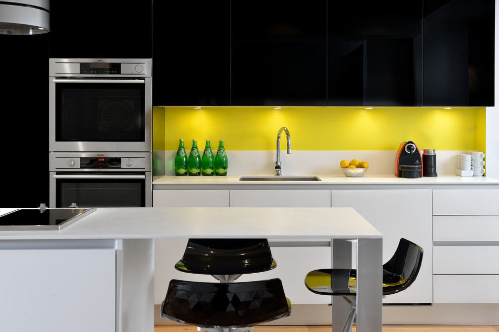 На фото: параллельная кухня-гостиная в стиле модернизм с врезной мойкой, плоскими фасадами, черными фасадами, столешницей из кварцита, желтым фартуком, техникой из нержавеющей стали, светлым паркетным полом и островом с