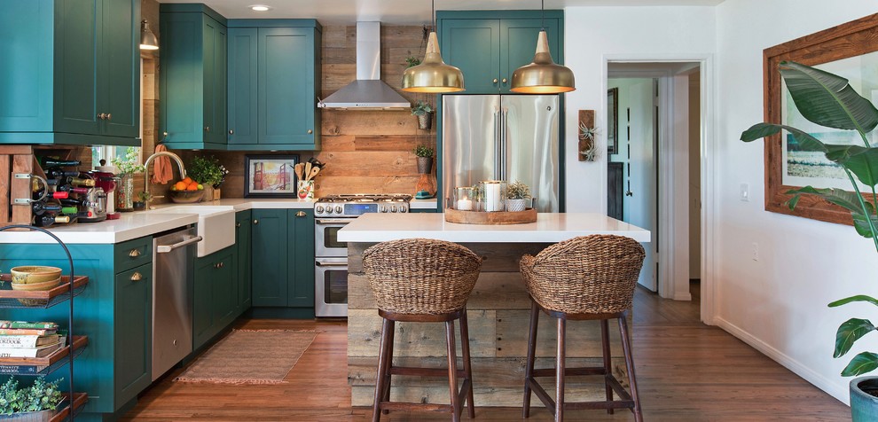 Example of a beach style l-shaped medium tone wood floor and brown floor kitchen design in San Diego with a farmhouse sink, shaker cabinets, turquoise cabinets, brown backsplash, wood backsplash, stainless steel appliances, an island and white countertops