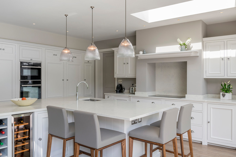This is an example of a large classic l-shaped kitchen/diner in Surrey with white cabinets, quartz worktops, grey splashback, laminate floors, an island, white worktops, marble splashback, a submerged sink, recessed-panel cabinets, integrated appliances and beige floors.