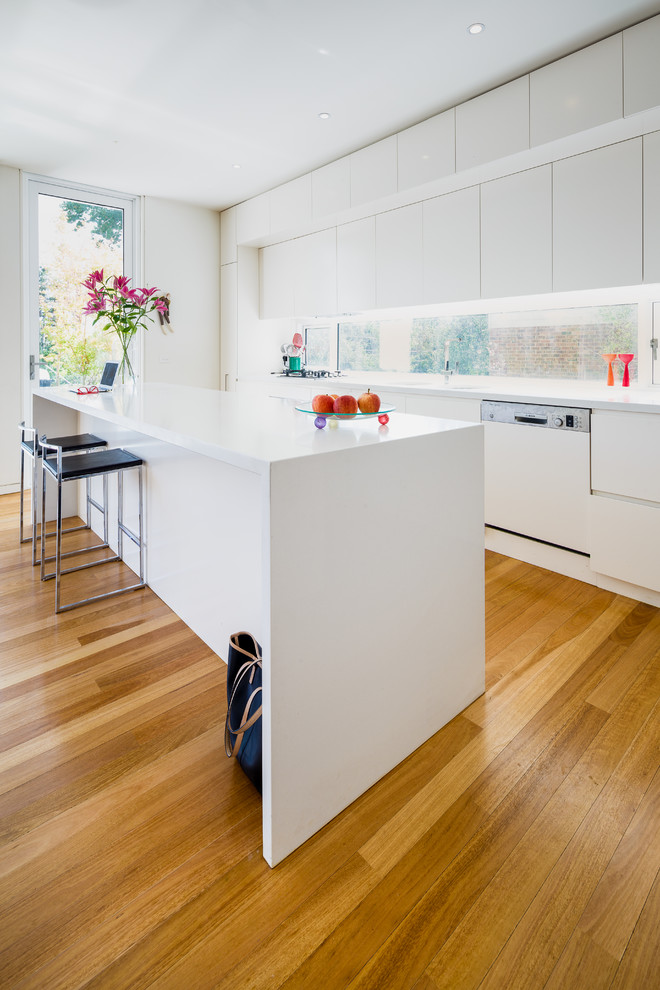 This is an example of a medium sized contemporary galley kitchen in Canberra - Queanbeyan with white cabinets, engineered stone countertops, window splashback, white appliances, medium hardwood flooring, an island, brown floors, white worktops and flat-panel cabinets.