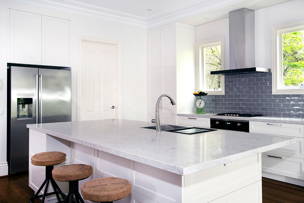 Inredning av ett klassiskt stort grå grått kök, med en dubbel diskho, skåp i shakerstil, vita skåp, bänkskiva i kvarts, grått stänkskydd, stänkskydd i tunnelbanekakel, rostfria vitvaror, mellanmörkt trägolv, en köksö och brunt golv