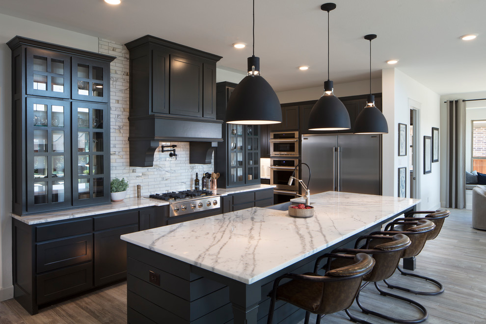 Example of a transitional l-shaped medium tone wood floor and brown floor kitchen design in Dallas with a farmhouse sink, recessed-panel cabinets, black cabinets, white backsplash, mosaic tile backsplash, stainless steel appliances, an island and white countertops