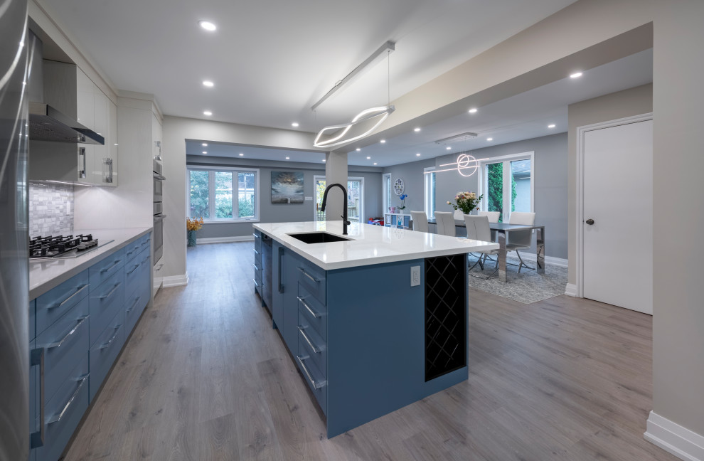 Modern inredning av ett stort linjärt kök och matrum, med en undermonterad diskho, skåp i shakerstil, blå skåp, grått stänkskydd, stänkskydd i keramik, rostfria vitvaror, ljust trägolv, en köksö och grått golv