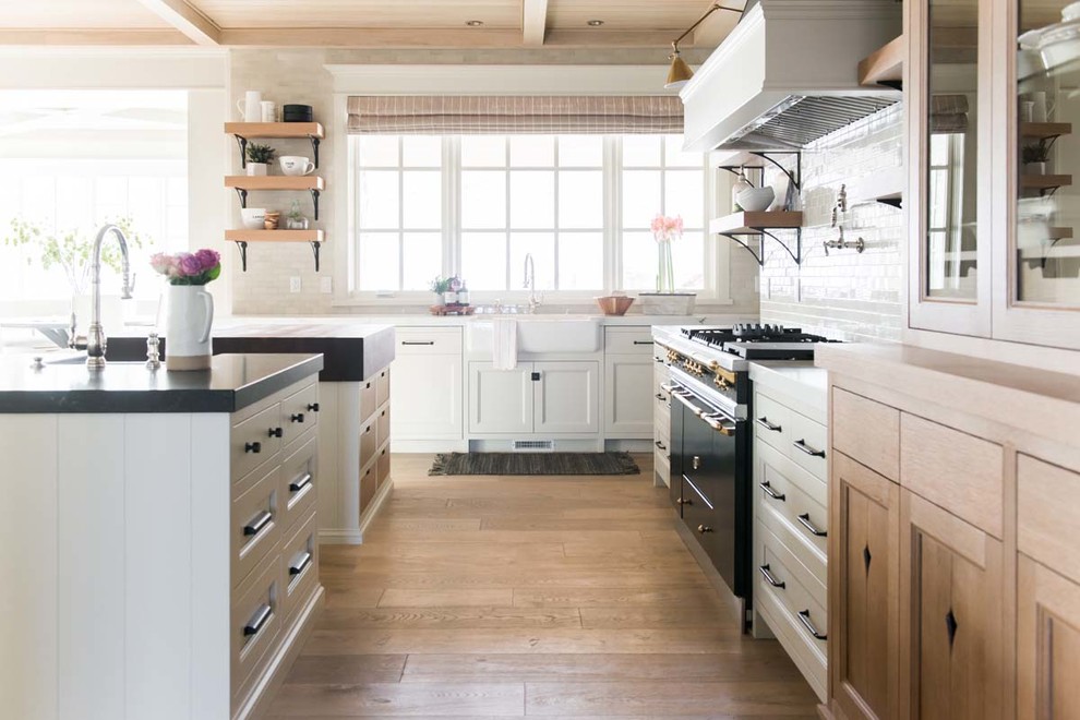 Bild på ett maritimt kök, med en rustik diskho, skåp i shakerstil, vita skåp, vitt stänkskydd, stänkskydd i tunnelbanekakel, svarta vitvaror, ljust trägolv, flera köksöar och beiget golv