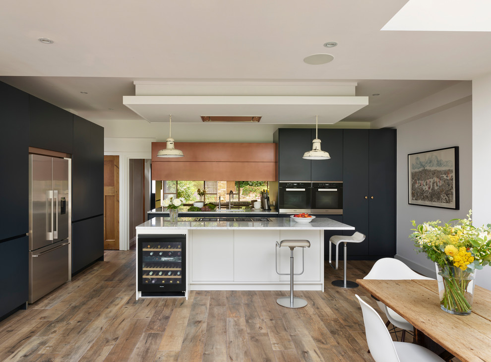 Large trendy l-shaped light wood floor and brown floor eat-in kitchen photo in London with an integrated sink, blue cabinets, marble countertops, metallic backsplash, mirror backsplash, an island and white countertops