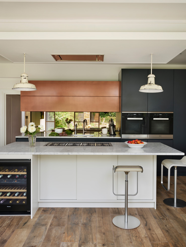 This is an example of a large contemporary l-shaped kitchen/diner in London with an integrated sink, blue cabinets, marble worktops, metallic splashback, mirror splashback, light hardwood flooring, an island, brown floors and white worktops.