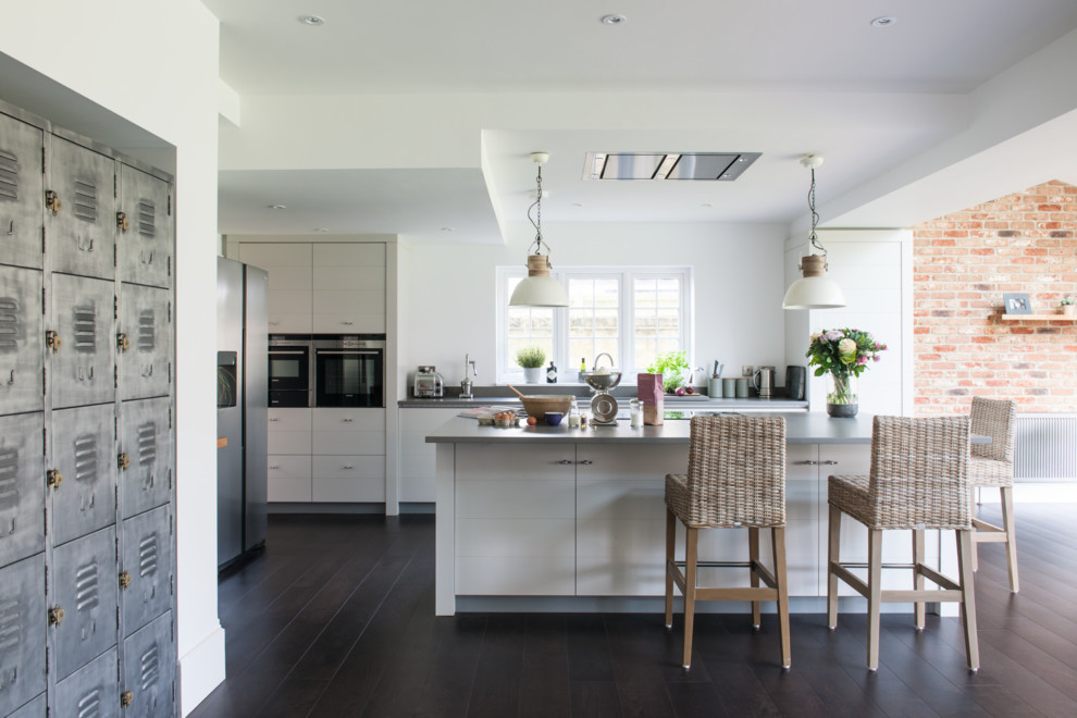 Inredning av ett lantligt stort grå linjärt grått kök, med släta luckor, vita skåp, rostfria vitvaror, mörkt trägolv, en köksö och brunt golv