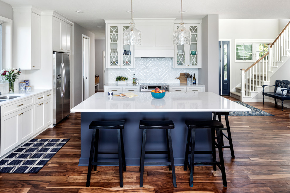 Inspiration for a medium sized coastal l-shaped open plan kitchen in Minneapolis with glass-front cabinets, white cabinets, engineered stone countertops, white splashback, ceramic splashback, stainless steel appliances, medium hardwood flooring, an island, white worktops and a submerged sink.